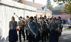 Eine Marschkapelle läuft auf der Straße, mehrere Menschen halten Musikinstrumente und tragen traditionelle Kleidung. Dahinter befindet sich ein Gebäude mit einer Flagge und Bäumen.