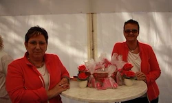 Zwei Frauen in roten Jacken stehen neben einem Tisch mit Blumen und einem Korb. Eine Frau hält eine Vase mit roten Blumen, die andere einen Korb mit einem Geschenk. Sie posieren für ein Foto.