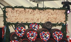 Ein Holzschild mit 'Iscvadorer' und einem Datum, umgeben von Fässern mit roten, weißen und blauen Kränzen, vor einem weißen Zelthintergrund.