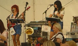 Drei Frauen spielen Instrumente auf einer Bühne vor einem Publikum. Zwei halten Gitarren, eine hat ein Keyboard, und eine Posaune steht in der Nähe. Es gibt Schilder und einen weißen Hintergrund.