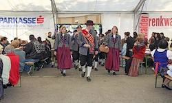 Eine Gruppe von Menschen in traditioneller Kleidung marschiert durch einen Zeltbereich, wobei Zuschauer auf Bänken sitzen, einige in roten und weißen Jacken. Im Hintergrund sind Banner sichtbar.