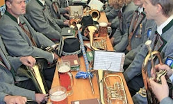 Eine Gruppe von Männern in grauen Uniformen sitzt um einen langen Tisch mit Musikinstrumenten, Gläsern und einem Buch. Die Instrumente umfassen Trompeten, einen Posaune und eine Flöte. Sie üben wahrscheinlich für eine Veranstaltung.