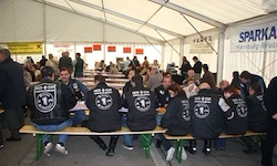 Eine Gruppe von Menschen in Jacken sitzt auf Bänken in einem Zelt. Sie essen. Das Zelt hat ein Banner mit der Aufschrift 'SPARKA.'