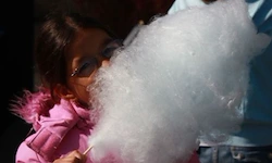Ein kleines Mädchen in einem rosa Mantel und mit Sonnenbrille bläst an einer großen Zuckerwatte.