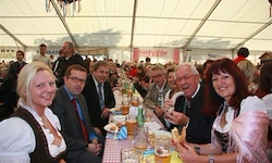 Eine Gruppe von Menschen, darunter Männer und Frauen, sitzen an einem Tisch in einem Zelt und genießen Essen und Getränke. Sie alle lächeln und scheinen eine gute Zeit zu haben.