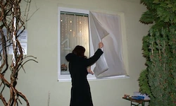 Eine Frau in einem schwarzen Mantel entfernt eine weiße Gardine von einem Fenster eines Gebäudes.
