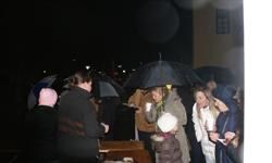 Eine Gruppe von Menschen steht im Regen und hält Regenschirme. Einige schauen auf einen Tisch mit Tassen, andere unterhalten sich. Das Gebiet ist dunkel.