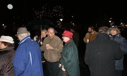 Eine Gruppe von Menschen steht im Dunkeln im Freien, einige mit Tassen. Sie tragen Winterkleidung und Hüte, einschließlich einer Frau mit einem roten Hut. Sie reden und halten einen Schirm.