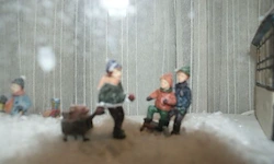 Eine Figur einer Frau und Kinder in Winterkleidung steht im Schnee in der Nähe eines Gebäudes. Sie halten einen roten Ball.