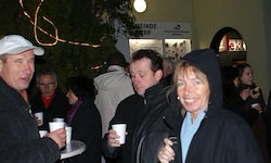 Eine Gruppe von Menschen steht bei Nacht draußen. Ein Mann und eine Frau lächeln und halten Tassen. Ein Mann trägt eine weiße Mütze. Die Menge ist in festlicher Stimmung.