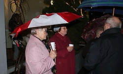 Zwei Frauen mit Regenschirmen, einer rot und einer weiß, stehen im Regen und halten Tassen. Sie sind von anderen umgeben, einige halten ebenfalls Regenschirme.