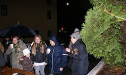 Eine Gruppe von Menschen steht zusammen, eingehüllt in Mäntel und Mützen, unter einem Regenschirm und in der Nähe eines Baumes, hält Tassen und unterhält sich bei Nacht.