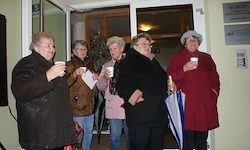 Fünf Frauen stehen in einer Tür, jede mit einer Tasse in der Hand. Sie scheinen zu plaudern. Eine Frau hält einen Regenschirm. Die Tür hat ein Schild mit dem Wort 'SCHORV'.