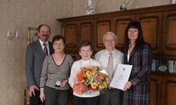 Fünf Menschen posieren für ein Foto in einem Raum. Eine Frau hält ein Zertifikat und einen Blumenstrauß. Hinter ihnen steht ein Holzschrank.