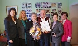Sechs Personen stehen eng beieinander und halten Blumen und ein Zertifikat. Die Person in der Mitte hält ein Zertifikat und Blumen. Ein gerahmtes Bild ist an der Wand hinter ihnen montiert.
