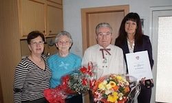 Vier Menschen stehen in einem Raum und halten Blumen und ein Zertifikat. Zwei Frauen und zwei Männer, wobei ein Mann einen Blumenstrauß und ein Zertifikat hält.