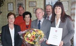 Eine Gruppe von Menschen posiert für ein Foto. Eine Frau hält ein Zertifikat und einen Blumenstrauß. Ein Mann im Anzug hält Blumen. Hinter ihnen hängen Bilderrahmen an der Wand.