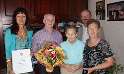 Fünf Menschen stehen eng beieinander und halten ein Zertifikat und Blumen. Die Frauen tragen Halsketten und Armbänder, eine mit einer Uhr. Sie stehen in einem Raum mit einem an der Wand montierten Bilderrahmen.