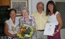 Vier Menschen posieren für ein Foto. Eine ältere Frau hält Blumen. Ein Mann hält ein Zertifikat. Eine Frau trägt eine Brille. Eine andere Frau hält ein Zertifikat. Ein Gemälde ist an der Wand.