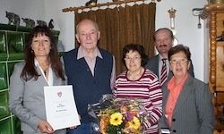 Eine Gruppe von Erwachsenen steht drinnen. Sie halten ein Zertifikat und einen Blumenstrauß. Eine Frau auf der linken Seite trägt eine Brille. Hinter ihnen ist eine Wand mit einem grünen Fliesenmuster und einem braunen Vorhang.