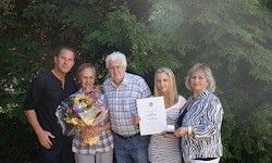 Fünf Menschen stehen zusammen in einem Garten und halten ein Zertifikat und Blumen. Der Mann links trägt ein schwarzes T-Shirt. Der Mann in der Mitte trägt ein kariertes Hemd. Die Frau rechts hält ein Zertifikat.