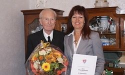 Ein älterer Mann und eine Frau stehen nebeneinander mit einem Blumenstrauß und einem Zertifikat in der Hand. Sie stehen vor einem Holzschrank mit verschiedenen Gegenständen darin.