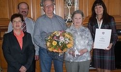 Fünf Menschen stehen drinnen zusammen. Die Frau in der Mitte hält einen Blumenstrauß. Die Frau auf der rechten Seite hält ein Zertifikat. Sie alle scheinen für ein Foto zu posieren.