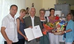 Eine Gruppe von Menschen posiert für ein Foto, wobei ein Mann ein Zertifikat und einen Blumenstrauß hält. Sie sind in einem Raum mit einem Kamin und einem Fenster.