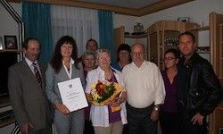 Eine Gruppe von Menschen steht drinnen zusammen, eine Frau hält einen Zertifikat und einen Blumenstrauß. Ein Mann in einer Lederjacke steht rechts.