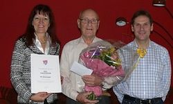 Drei Personen stehen nebeneinander. Eine Frau hält ein Zertifikat und ein Mann eine Blumenbouquet. Sie scheinen einen besonderen Anlass zu feiern.