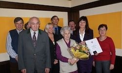 Eine Gruppe von Menschen steht zusammen und hält Blumen und ein Zertifikat. Eine ältere Frau hält die Blumen und das Zertifikat. Sie befinden sich in einem Raum mit gelben und weißen Wänden.