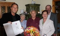 Fünf Menschen posieren drinnen, ein Mann hält ein Papier, ein anderer hält Blumen, drei tragen formelle Kleidung, ein grüner Schrank und eine weiße Statue im Hintergrund.