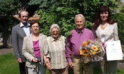 Fünf Personen stehen in einem Garten mit Bäumen und Rasen. Eine Frau hält Blumen und eine andere ein Zertifikat. Sie lächeln und posieren für ein Foto.