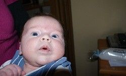 Ein Neugeborenes mit blauen Augen, das einen blau-weiß gestreiften Stoff trägt, wird von einer Person in einem Raum gehalten.