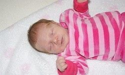 Ein Babymädchen schläft mit geschlossenen Augen auf einer rosa Decke. Sie trägt rosa und weiße gestreifte Schlafanzughosen.