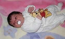Ein Baby in einem Winnie-the-Pooh-Overall liegt auf einer Decke mit blauen und rosa Designs. Das Baby saugt an einem Schnuller.
