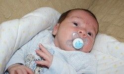 Ein Baby mit einem Schnuller liegt auf einer gelben Decke mit weißen Punkten und schaut mit leicht geöffnetem Mund in die Kamera.