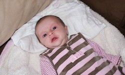 Ein Baby in rosa und braun gestreifter Kleidung liegt auf einer weißen Decke und schaut mit großen Augen nach oben.