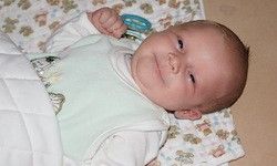 Ein Baby liegt auf einem Bett mit einem blauen Spielzeug in der Hand, trägt ein grünes Lätzchen und weißen Stoff, lächelt und schaut in die Kamera.