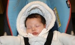 Ein Baby schläft gemütlich in einem Autositz, eingewickelt in einen weißen Wintermantel mit Kapuze, und zeigt einen friedlichen Ausdruck auf seinem Gesicht.