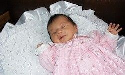 Ein kleines Mädchen in einem rosa Kleid schläft friedlich auf einem weißen Tuch, die Hände erhoben. Sie ruht auf einem Bett mit einer weißen Decke.