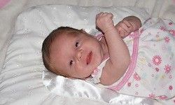 Ein Babymädchen liegt auf einer weißen Decke mit Blumenmuster und trägt ein rosa-weißes Kleid. Sie schaut direkt in die Kamera, die Hände erhoben und lächelt.