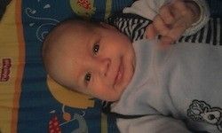 Ein lächelndes Baby liegt auf einer blauen Decke mit verschiedenen bunten Mustern. Das Baby trägt ein hellblaues T-Shirt mit einem Comic-Charakter darauf.