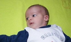 Ein Baby liegt auf einer grünen Decke und trägt ein weißes Shirt mit blauen Ärmeln und dem Wort 'DACH' darauf. Das Baby schaut direkt in die Kamera.