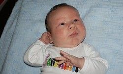 Ein Baby liegt auf einer blauen Decke und trägt ein weißes Shirt mit bunten Buchstaben und einem Regenbogendesign. Die Augen des Babys sind geöffnet und es scheint etwas anzuschauen.