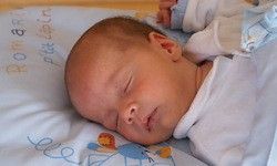 Ein Babyjunge schläft auf einer blau-weißen Decke mit musikalischen Noten und einem Löwendesign. Er hat einen Schnuller im Mund und einen kleinen blauen Teddybär in der Nähe.