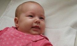 Ein lächelndes Baby liegt auf einem weißen Tuch und trägt ein rosa Oberteil mit kleinen weißen Mustern. Die Augen des Babys sind offen und es scheint etwas anzuschauen.