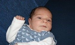 Ein Neugeborenes liegt auf einer blauen Oberfläche und trägt einen blau-weißen Strampler mit einem weißen langen Ärmel. Das Baby hat einen ernsten Ausdruck und eine Hand ist leicht erhoben.