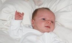 Ein Baby liegt auf einer weißen Decke und scheint für ein Foto zu posieren. Es trägt ein weißes Outfit und hebt eine Hand.