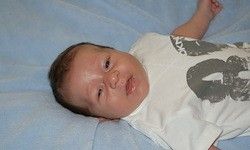 Ein Baby liegt auf einer blauen Decke mit einem weißen T-Shirt, auf dem eine graue 8 abgebildet ist. Das Baby hat braune Haare und einen kleinen Mund.